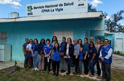 Grupo de trabajadores de salud del centro de atención primaria La Vigía y expertos de OPS posan frente al centro de salud, que tiene las paredes pintadas de verde aguamarina, con un cielo azul con nubes dispersas y un frente de hierba